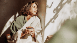 a woman with linen blankets draped around her holds a mug of tea and looks out a sunny window