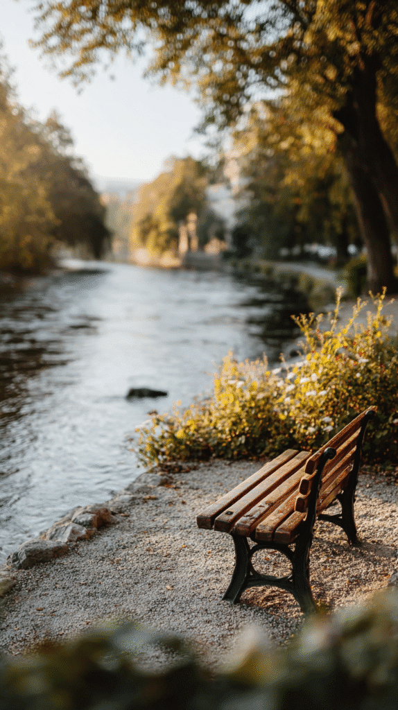 Empty park bench beside a serene river, surrounded by greenery, under a warm sunlight glow.