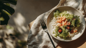 Plate of rice with fresh salad, cherry tomatoes, and salmon on a wooden table with fork and linen near a plant.