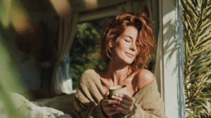 Woman enjoying sunlight by the window with a cup of coffee, wrapped in a cozy sweater.